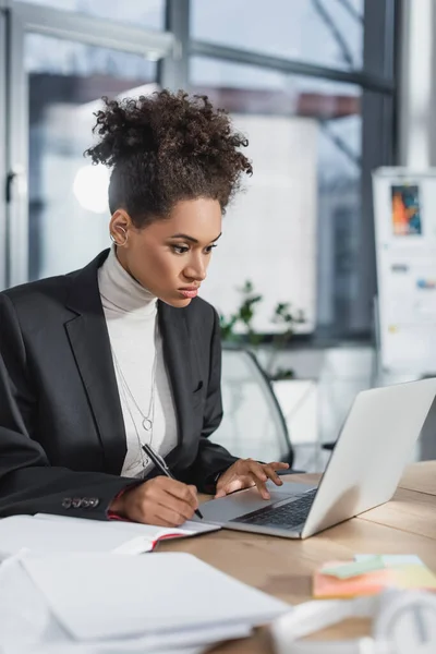 Empresária afro-americana usando laptop e escrevendo no notebook no escritório — Fotografia de Stock