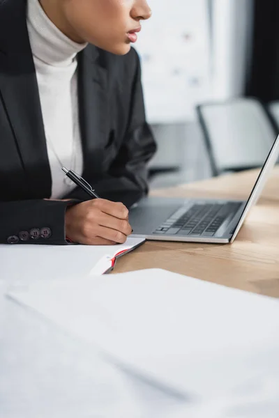 Ausgeschnittene Ansicht einer afrikanisch-amerikanischen Geschäftsfrau, die in der Nähe eines Laptops im Büro auf einem Notizbuch schreibt — Stockfoto