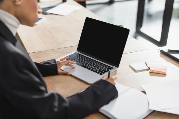 Ausgeschnittene Ansicht einer verschwommenen afrikanisch-amerikanischen Geschäftsfrau, die im Büro Laptop benutzt und auf ein Notizbuch schreibt — Stockfoto