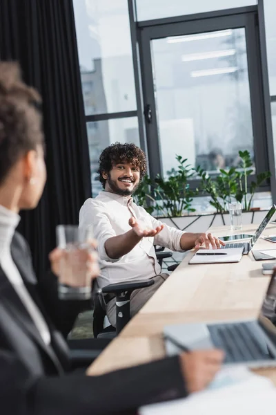 Positive indian businessman pointing with hand near devices and blurred african american colleague in office — Stock Photo