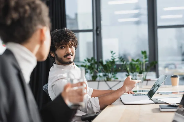 Indischer Geschäftsmann zeigt auf Laptop neben verschwommenem afrikanisch-amerikanischem Kollegen mit Wasser im Büro — Stockfoto