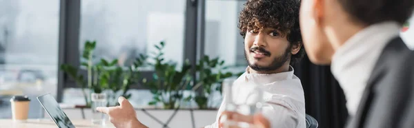 Uomo d'affari indiano che indica un portatile vicino a un collega afroamericano sfocato in carica, striscione — Foto stock