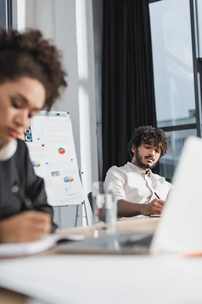 Junger indischer Geschäftsmann hält Stift neben verschwommenem afrikanisch-amerikanischem Kollegen und Flipchart im Büro — Stockfoto