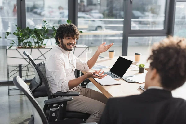 Hombre de negocios indio sonriente hablando con un borroso colega afroamericano cerca de la computadora portátil y el cuaderno en la oficina - foto de stock