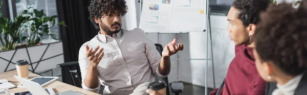 Empresario indio hablando con colegas afroamericanos cerca del café para llevar y dispositivos en la oficina, pancarta - foto de stock