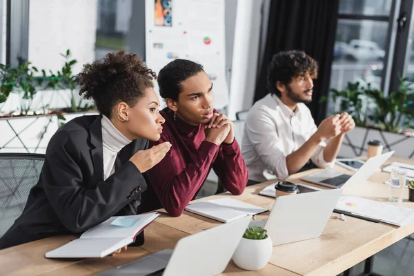 Empresarios afroamericanos hablando cerca de computadoras portátiles y colega indio borroso en la oficina - foto de stock