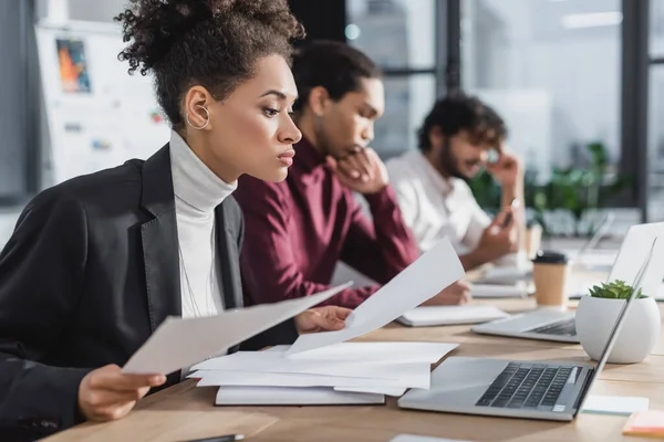 Jeune femme d'affaires afro-américaine tenant des papiers près d'un ordinateur portable au bureau — Photo de stock