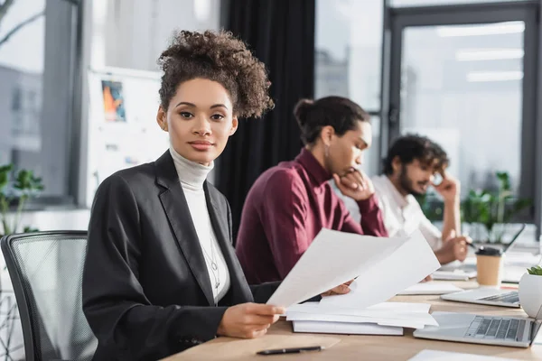 Junge afrikanisch-amerikanische Geschäftsfrau blickt in die Kamera in der Nähe von Laptop und verschwommenen Geschäftsleuten im Büro — Stockfoto