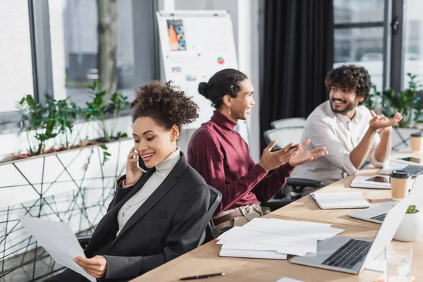 Empresária afro-americana positiva conversando no smartphone e segurando papel perto de colegas inter-raciais borrados no escritório — Fotografia de Stock