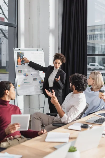 Africano americano empresária apontando para flip chart perto de colegas inter-raciais durante reunião no escritório — Fotografia de Stock