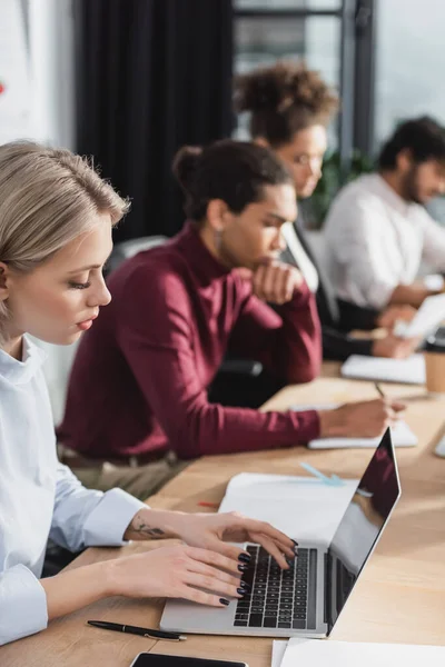 Joven empresaria que usa portátil con tableta digital cerca de colegas multiétnicos borrosos en la oficina - foto de stock