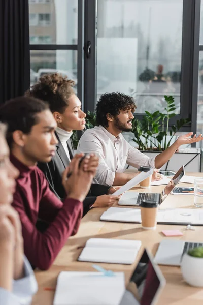 Seitenansicht eines indischen Geschäftsmannes, der in der Nähe von Geräten spricht, und verschwommener multiethnischer Kollegen bei einem Treffen im Büro — Stockfoto