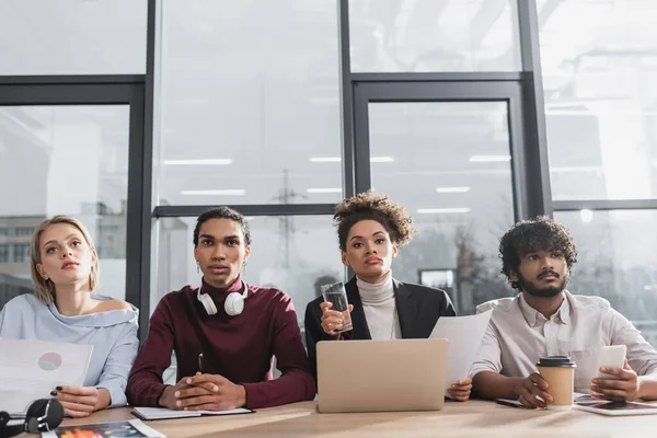 Jóvenes empresarios multiculturales sentados cerca de dispositivos y papeles en la oficina - foto de stock