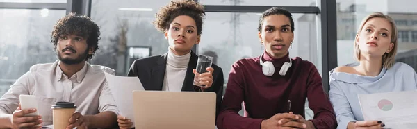 Interrassische Geschäftsleute, die in der Nähe von Zeitungen, Laptop und Kaffee im Büro wegschauen, Banner — Stockfoto