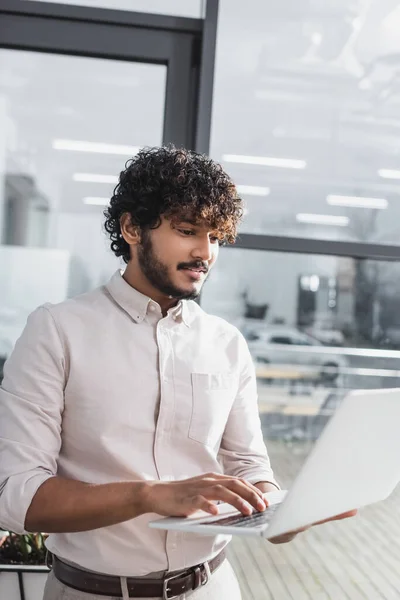 Empresário indiano sorridente usando laptop desfocado no escritório — Fotografia de Stock