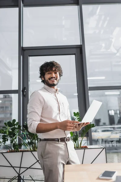 Positiver indischer Geschäftsmann blickt in Kamera und hält Laptop im Büro — Stockfoto