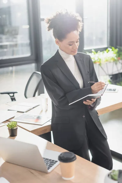 Afroamerikanische Geschäftsfrau schreibt auf Notizbuch neben verschwommenem Laptop und Coffee to go im Büro — Stockfoto