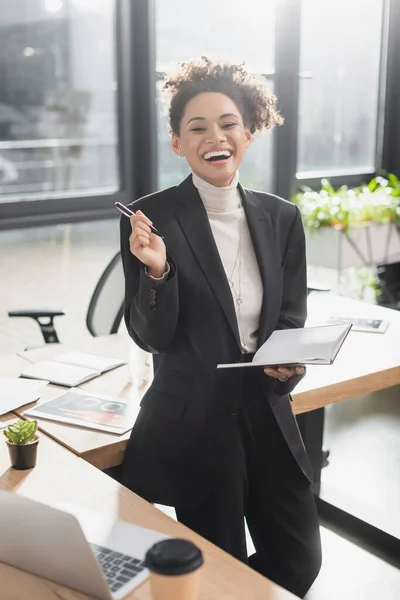 Alegre mujer de negocios afroamericana sosteniendo un cuaderno y mirando a la cámara en la oficina - foto de stock