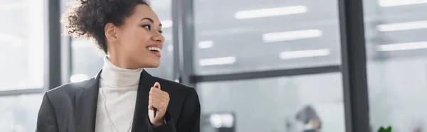 Mujer de negocios afroamericana sonriente sosteniendo la pluma en la oficina, pancarta - foto de stock