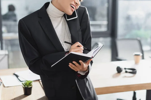 Cropped view of african american businesswoman talking on smartphone and holding notebook in office — Stock Photo