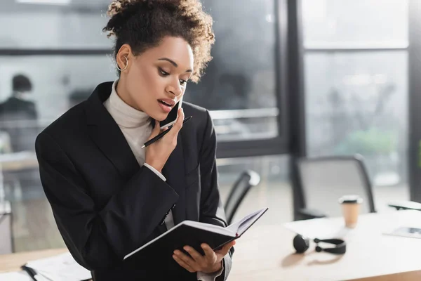 Mujer de negocios afroamericana mirando el cuaderno mientras habla en el teléfono inteligente en la oficina - foto de stock