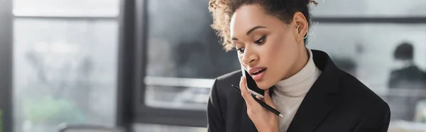 Joven mujer de negocios afroamericana sosteniendo pluma y hablando por teléfono celular en la oficina, pancarta - foto de stock