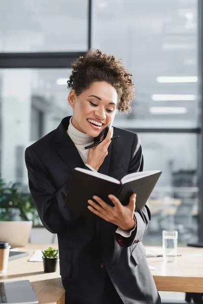 Empresária afro-americana positiva segurando notebook e falando no smartphone no escritório — Fotografia de Stock