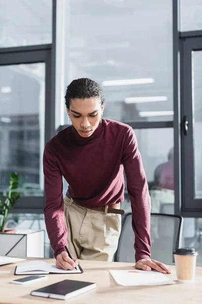 Afrikanischer Geschäftsmann schreibt auf Notizbuch in der Nähe von Smartphone und Coffee to go im Büro — Stockfoto