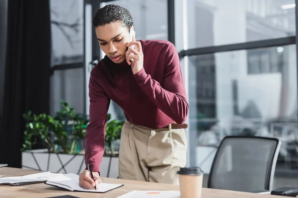 Hombre de negocios afroamericano hablando por teléfono inteligente y escribiendo en un cuaderno en la oficina - foto de stock