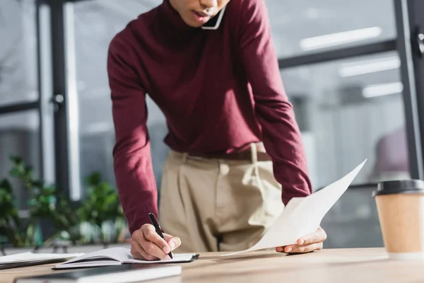 Vista ritagliata dell'uomo d'affari afro-americano che scrive sul taccuino e tiene la carta mentre parla al cellulare in ufficio — Foto stock