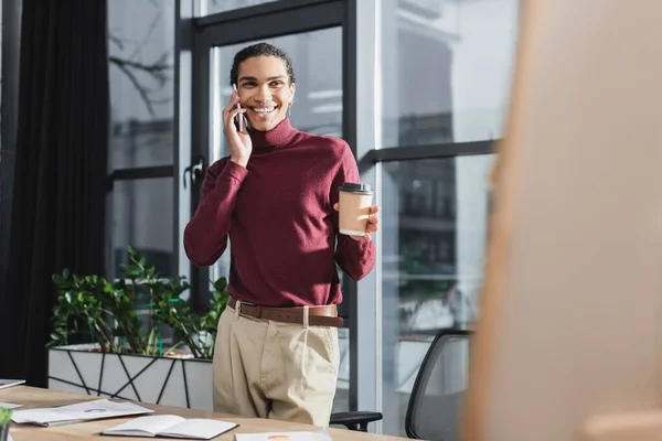 Sonriente hombre de negocios afroamericano hablando en el teléfono inteligente y sosteniendo café para ir cerca de portátil en la oficina - foto de stock