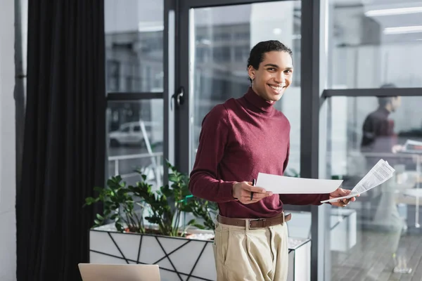 Улыбающийся африканский американский бизнесмен с бумагами возле ноутбука в офисе — стоковое фото