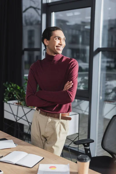 Улыбающийся африканский американский бизнесмен, стоящий рядом с кофе, чтобы пойти и ноутбук в офисе — стоковое фото