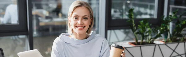 Positive Geschäftsfrau mit Coffee to go und digitalem Tablet mit Blick auf Kamera im Büro, Banner — Stockfoto