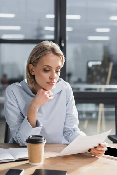 Joven empresaria mirando documentos cerca de dispositivos y café en la oficina - foto de stock