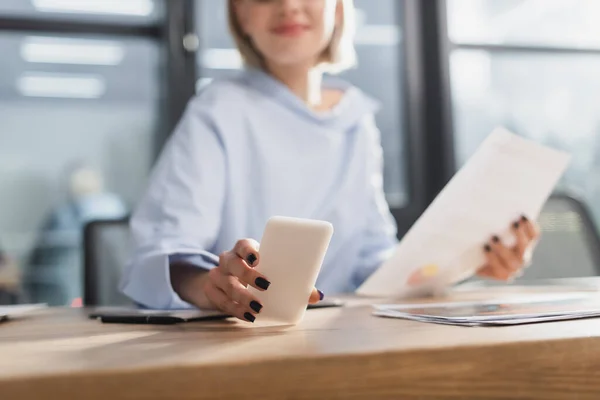 Vista recortada de una mujer de negocios borrosa usando un teléfono inteligente cerca de los papeles en la oficina - foto de stock