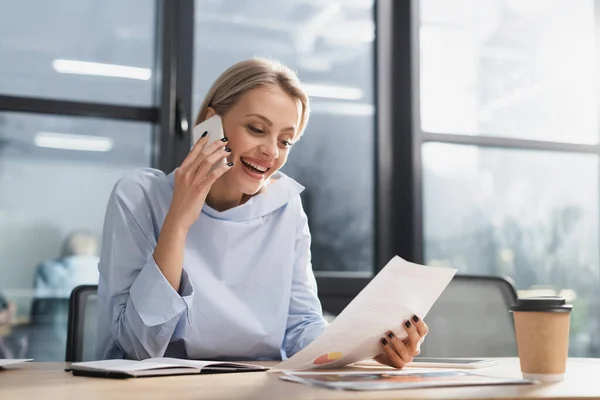 Cheerful businesswoman talking on smartphone and holding paper near coffee to go in office — Stock Photo