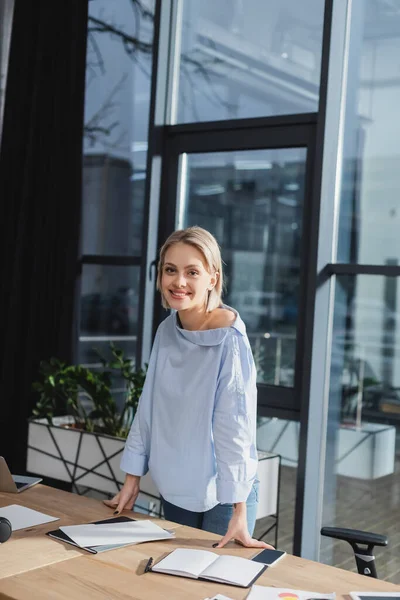 Femme d'affaires positive regardant la caméra près des documents et des appareils dans le bureau — Photo de stock