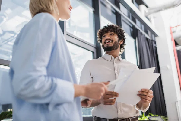 Tiefansicht eines fröhlichen indischen Geschäftsmannes, der Dokumente in der Nähe eines verschwommenen Kollegen im Amt hält — Stockfoto