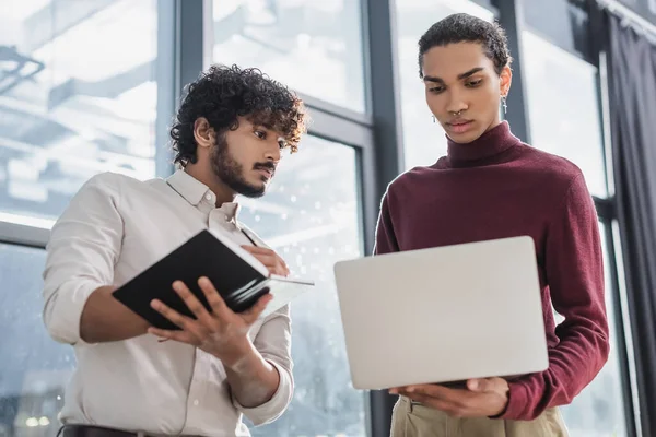 Uomo d'affari indiano che scrive su taccuino vicino a collega afro-americano con laptop in carica — Foto stock