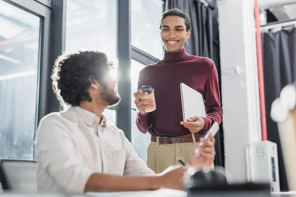 African american businessman holding coffee to go and laptop near indian colleague in office — Stock Photo