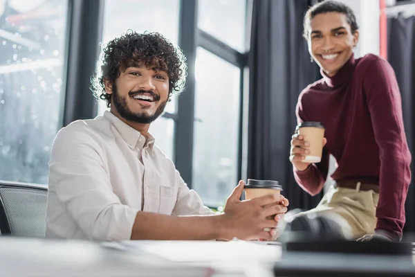 Cheerful indian businessman holding coffee to go near blurred african american colleague in office — Stock Photo