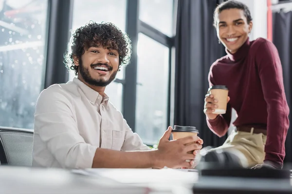 Fröhlicher indischer Geschäftsmann hält Kaffee in der Nähe eines verschwommenen afrikanisch-amerikanischen Amtskollegen — Stockfoto