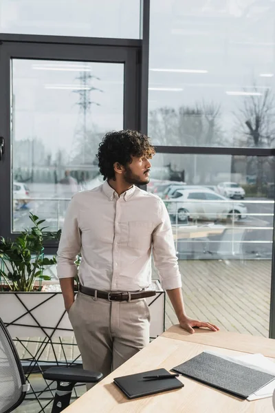 Vista laterale del giovane uomo d'affari indiano in piedi vicino al tavolo da lavoro in ufficio — Foto stock