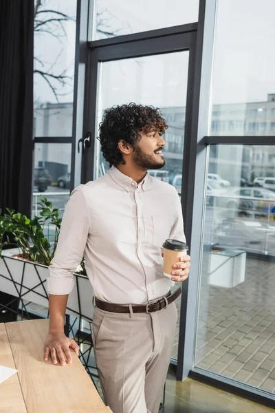 Side view of positive indian businessman holding coffee to go in office — Stock Photo