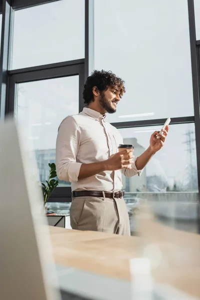 Vista lateral de un empresario indio sonriente sosteniendo café para llevar y usando un teléfono inteligente en la oficina - foto de stock