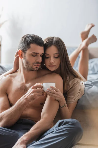 Mulher bonita segurando xícara de café perto de namorado sem camisa no quarto — Fotografia de Stock