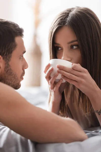 Morena mulher bebendo café e olhando para o namorado borrado no quarto — Fotografia de Stock