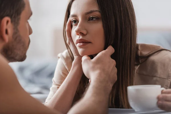 Blurred man touching face of brunette girlfriend with cup of coffee in bedroom — Stock Photo