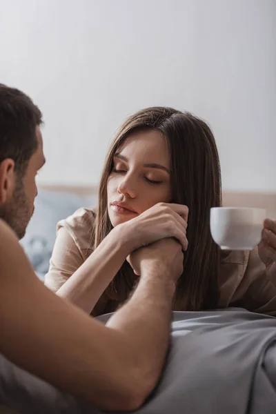 Blurred man holding hand of pretty girlfriend with coffee cup on bed — Stock Photo